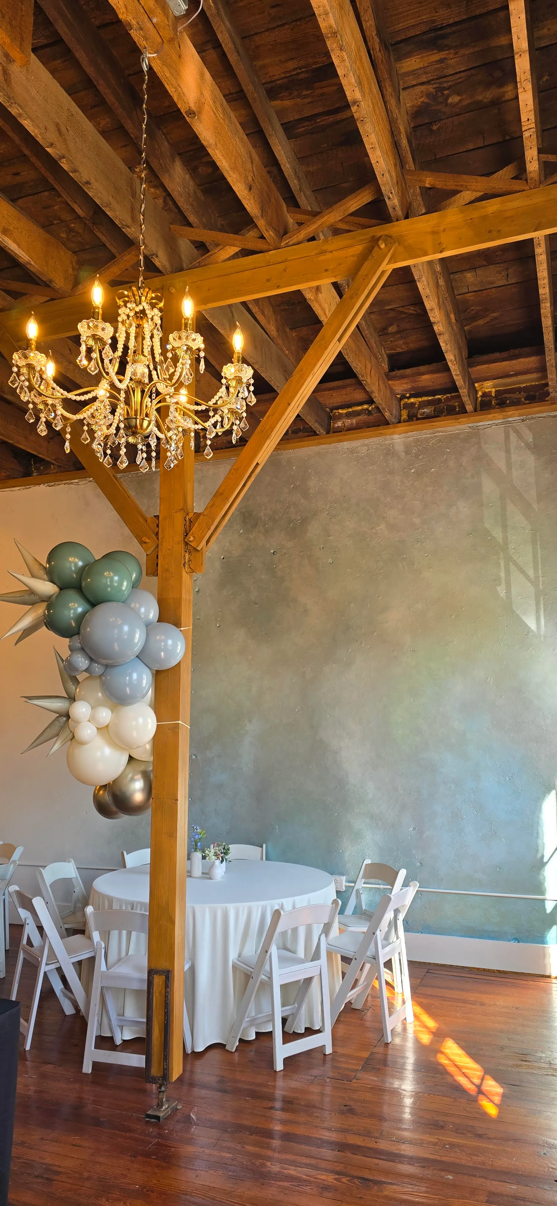 Event reception space with round table setup, white chairs, chandelier, exposed wood beams, and balloon decor